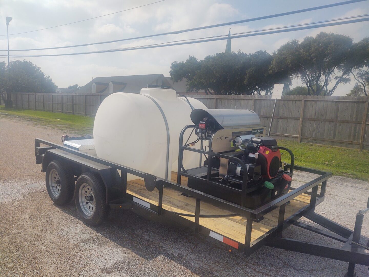 An industrial cleaning equipment water tank, white in color, mounted on a black dual-axle trailer with a generator and pump, parked on a gravel lot under clear skies.
